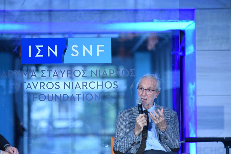 Ένας άντρας κάθεται στη σκηνή και μιλάει κρατώντας ένα μικρόφωνο. Από πίσω του υπάρχει ένα μπλε φόντο που γράφει ΙΔΡΥΜΑ ΣΤΑΥΡΟΣ ΝΙΑΡΧΟΣ STAVROS NIARCHOS FOUNDATION 