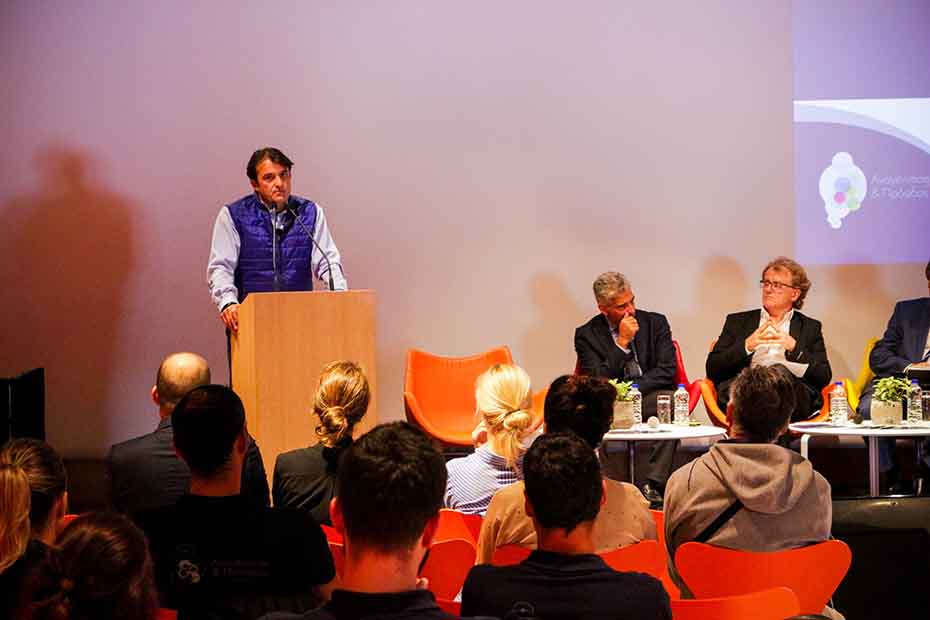 Ένας άντρας μιλάει στο βήμα ενώ δίπλα του στη σκηνή κάθονται και άλλοι άντρες και το κοινό από κάτω τον παρακολουθεί 