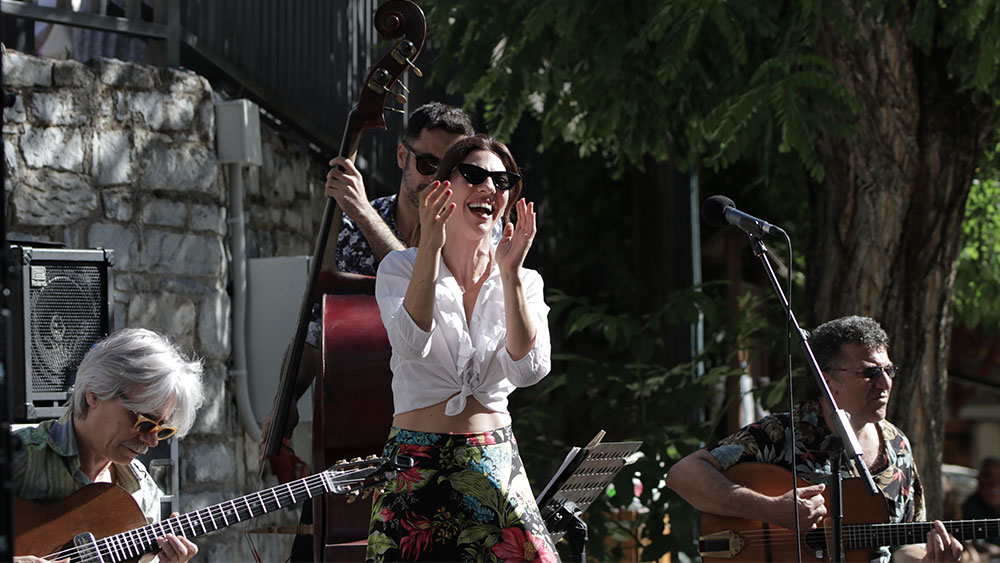 Μια γυναίκα στέκεται όρθια μπροστά από το μικρόφωνο γελώντας και χτυπώντας παλαμάκια και πίσω της είναι τρεις άντρες με μουσικά όργανα 